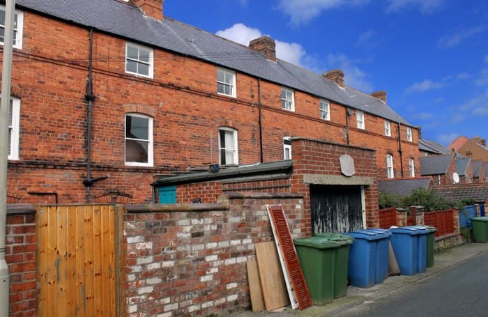 A typical residential street features red brick terraced houses and numerous green and blue recycling bins along the pavement with some discarded household waste items leaning against a low brick wall illustrating the importance of proper waste disposal to prevent fly-tipping.